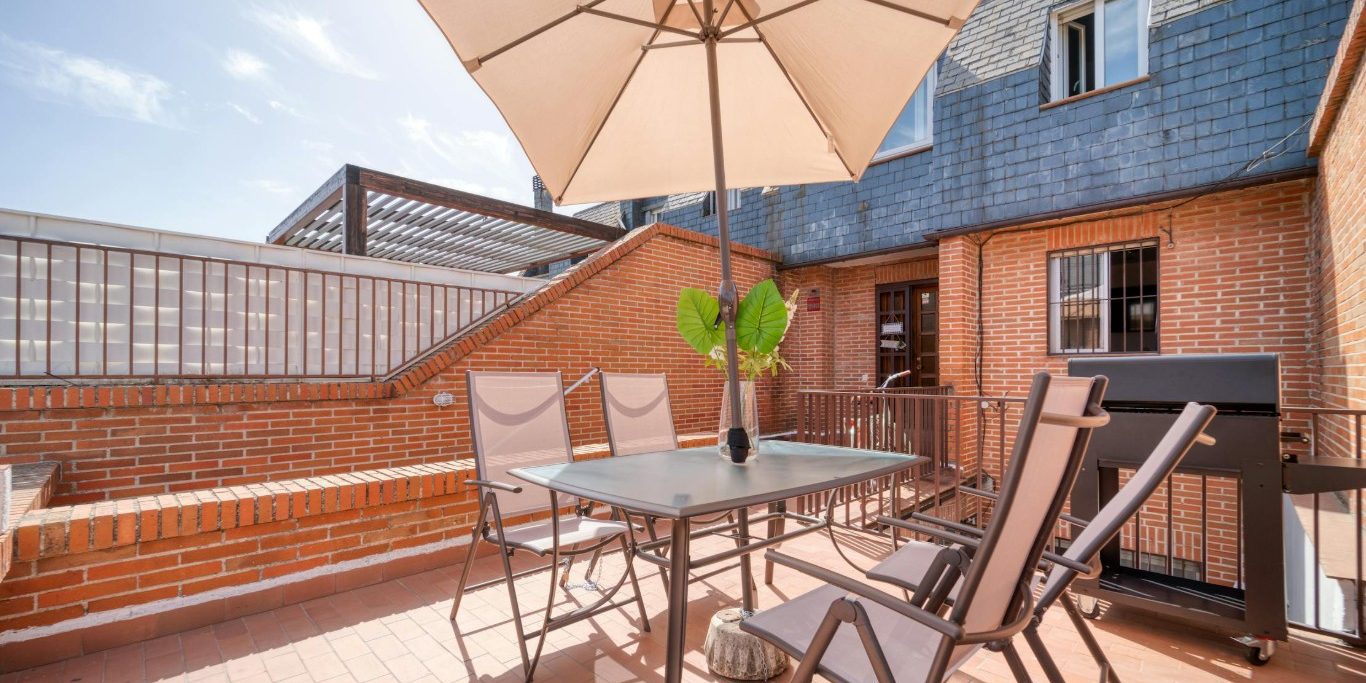 Zonnig dakterras met metalen tafel, vier stoelen, parasol en barbecue tegen een gevel met bakstenen