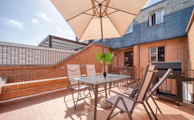 Zonnig dakterras met metalen tafel, vier stoelen, parasol en barbecue tegen een gevel met bakstenen